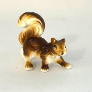 May include: A ceramic squirrel figurine with a brown body, a cream-colored belly and paws, and a large, curved tail. The squirrel is posed with one paw extended, giving the impression of movement. The figurine has a glossy finish.