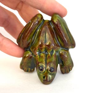 May include: A ceramic frog figurine with a glossy finish. The frog is primarily green with brown accents and black eyes. The figurine has a detailed design, including the frog's legs and face. The frog is a decorative item.