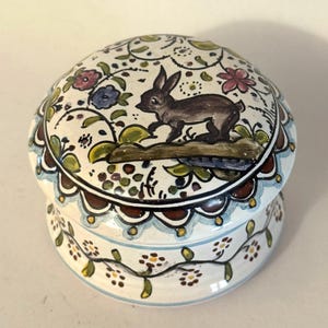 May include: A round, ceramic trinket box with a lid. The box is white with a painted design of a brown rabbit, flowers, and vines. The rim of the lid and base are decorated with a scalloped pattern in blue and brown.