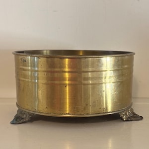 Puede incluir: Un cuenco redondo de latón con un acabado metálico pulido y detalles de ranuras horizontales a lo largo de la pared exterior. El recipiente se apoya sobre tres pies de metal plateado ornamentados con intrincados patrones decorativos de hojas.