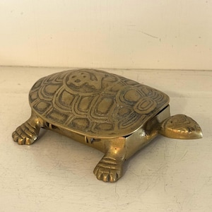 Könnte beinhalten: Eine messingfarbene Schmuckschatulle in Schildkrötenform. Die Schatulle hat ein detailliertes Schalenmuster und wird von vier Füßen getragen. Der Kopf der Schildkröte ragt nach vorne, und die Schatulle öffnet sich oben. Das Messing hat eine warme, gealterte Patina.