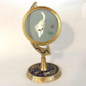 May include: A decorative desk mirror with a round, gold-toned frame. The mirror features an embroidered image of a white cat with black eyes and a green insect on a light blue background. The base is gold with a blue and gold dragon design.