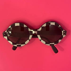 May include: Round sunglasses with a black and white checkered pattern on the frames. The sunglasses have dark lenses and black arms. The background is a bright pink.