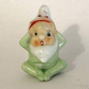 Könnte beinhalten: Kleine Keramik-Wichtelfigur in sitzender Pose, die Hände an den Ohren. Der Wichtel trägt ein hellgrünes Outfit, einen weißen Bart und einen weißen Hut mit rotem Band. Die Figur hat rosige Wangen und einen überraschten Gesichtsausdruck.