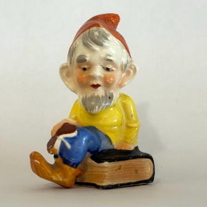 Könnte beinhalten: Keramik-Wichtelfigur mit roter Mütze, gelbem Hemd, blauer Hose und beigen Stiefeln. Der Wichtel hat einen weißen Bart und sitzt auf einem Buch. Die Figur ist glänzend lackiert.