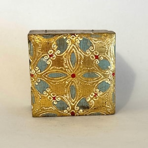 May include: A square, decorative box with a gold and teal floral pattern. The gold design features intricate details and red accents. The box has a wooden appearance and is likely used for storage or display.