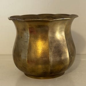 Puede incluir: Una maceta de metal con acabado en tono latón y borde festoneado. El recipiente presenta un cuerpo redondeado y bulboso con paneles verticales acanalados que crean una superficie texturizada. El metal muestra una pátina desgastada con reflejos.