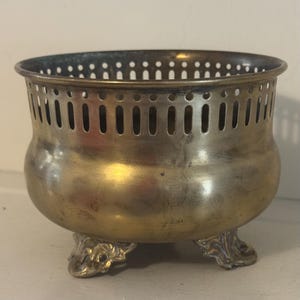 May include: A round brass bowl with a polished finish and a wide, open top. The upper rim features a decorative gallery with vertical cutouts. The bowl is supported by three ornate, curved feet that provide elevation and stability to the structure.
