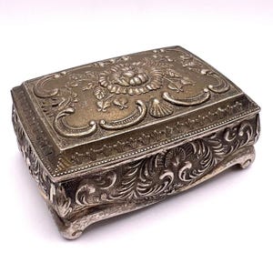 Puede incluir: Una caja de joyería rectangular plateada con detalles florales y de volutas ornamentados. La tapa presenta un diseño floral central rodeado de hojas y adornos curvos. La caja descansa sobre pequeños pies curvos.