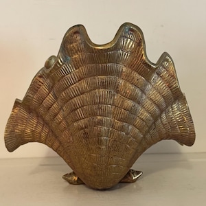 May include: A brass shell-shaped decorative object featuring a textured, fan-like surface with radiating vertical ridges. The metallic piece has a scalloped top edge and a wide, curved form that tapers toward a small, stable base at the bottom.