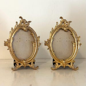 May include: Two ornate, gold-colored oval picture frames. Each frame features an elaborate design with a small cherub at the top and a decorative base. The frames are displayed on a white surface.