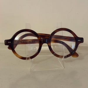 May include: Round tortoiseshell eyeglasses with clear lenses. The frames have a dark brown and amber pattern. The temples feature star-shaped accents. The text "N ROBERTS LA 1 3322" is visible on the frame.