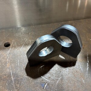 Könnte beinhalten: Zwei Metallhalterungen im Industriestil mit abgerundeten Ecken und einem zentralen kreisförmigen Loch. Die Halterungen bestehen aus dunklem, unlackiertem Metall und liegen auf einer strukturierten Metalloberfläche. Die Beleuchtung erzeugt Schatten.