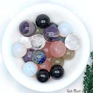 May include: A collection of polished crystal spheres in various colors, including clear, pink, purple, blue, and black, displayed in a white bowl.