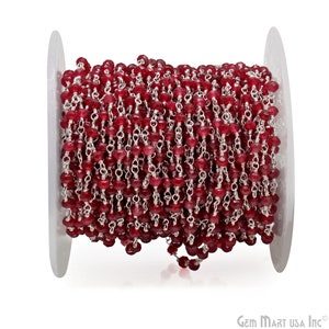 May include: A spool of red gemstone beads strung on a silver chain. The chain is made of small, linked silver rings. The beads are faceted and have a shiny finish.