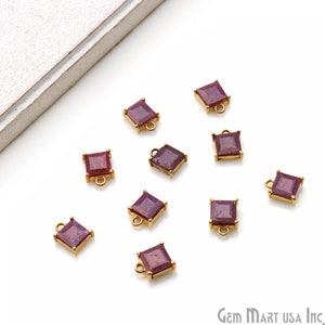 May include: Ten square-shaped ruby red gemstone charms with gold-toned metal loops. The charms are scattered on a white surface.