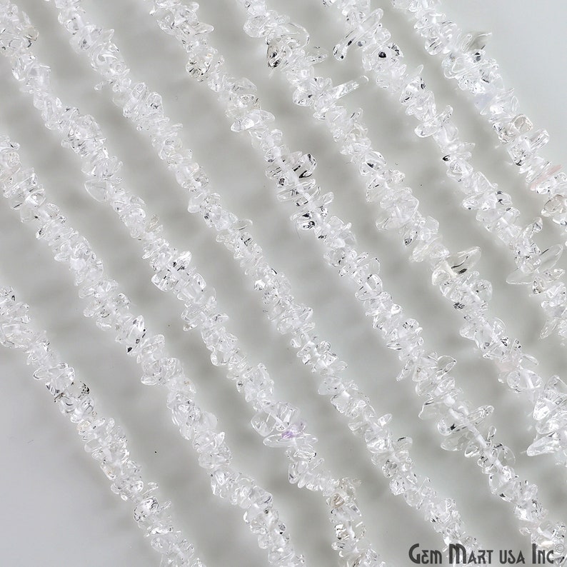 May include: A strand of clear quartz crystal chips, also known as rock crystal, arranged in parallel lines on a white background. The chips are irregular in shape and have a translucent appearance. Gem Mart USA Inc. is printed in the bottom right corner of the image.