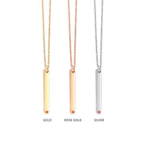 May include: Three gold bar necklaces with a small red gemstone at the bottom of the bar. The necklaces are shown in gold, rose gold, and silver.