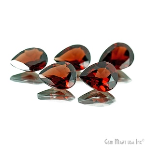 May include: Four pear-shaped red gemstones, likely garnets, are arranged on a white surface. The gemstones are faceted and reflect light.