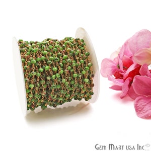 May include: A spool of green and gold rosary chain with small round beads. The chain is wound around a white spool and is next to a pink flower.