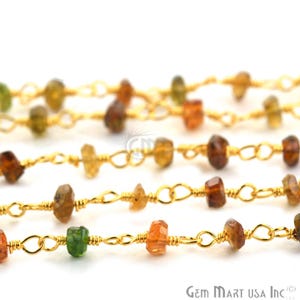 May include: A gold-toned metal chain featuring small, irregularly shaped, multi-coloured gemstone beads in shades of green, amber, and brown. The beads are individually wire-wrapped and spaced along the delicate links of the chain.