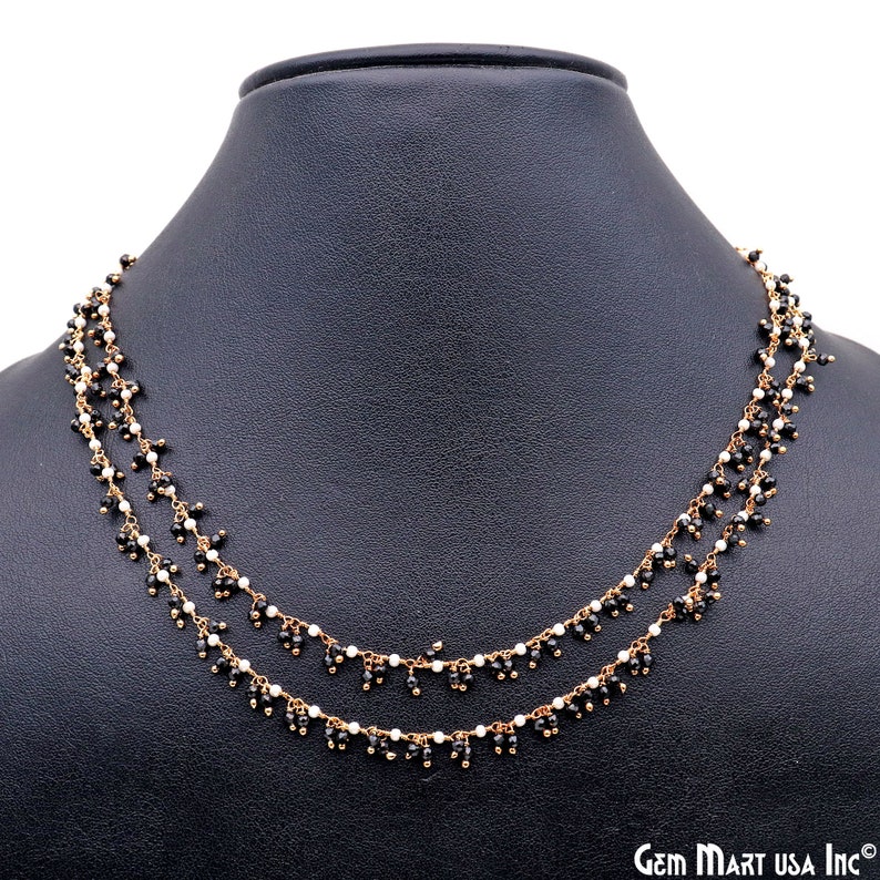 K&ouml;nnte beinhalten: Eine zweireihige Halskette mit kleinen schwarzen und wei&szlig;en Perlen auf einer goldfarbenen Kette. Die Halskette wird auf einer schwarzen Schaufensterpuppe pr&auml;sentiert. Das Logo von Gem Mart USA Inc. ist unten rechts sichtbar.