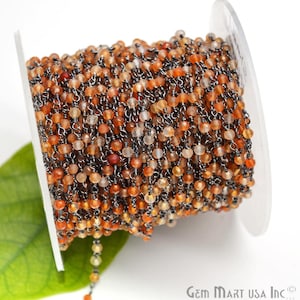 May include: A spool of orange carnelian gemstone beads strung on a silver chain. The beads are small and round, and the chain is delicate. The spool is white and has a label that says "Gem Mart USA Inc."