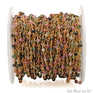 May include: A spool of gold chain with multi-colored gemstone beads. The beads are a mix of pink, green, and black. The chain is made of gold-plated metal.