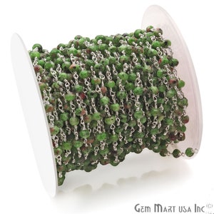 May include: A spool of green and red gemstone beads strung on a silver chain. The beads are faceted and have a natural, rough appearance. The chain is delicate and has a simple design.