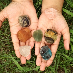 May include: A collection of seven different colored crystals, including green, blue, pink, white, brown, and yellow, are displayed in the palms of a person's hands.