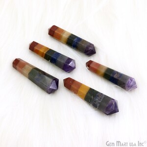 Pode incluir: Cinco varinhas de cristal de sete chakras multicoloridas, cada uma com uma ponta pontiaguda, dispostas sobre uma superfície branca e felpuda. As varinhas são feitas de sete pedras de diferentes cores, incluindo vermelho, laranja, amarelo, verde, azul, índigo e violeta.