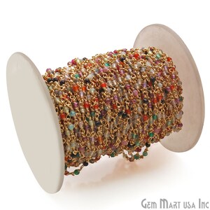 May include: A spool of gold chain with colorful gemstone beads. The chain is made of small, linked gold rings and is interspersed with tiny, faceted gemstones in various colors, including red, green, blue, and purple.