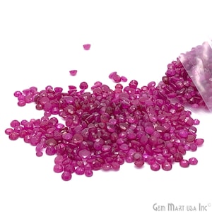 May include: A large pile of loose ruby red gemstones, scattered on a white surface. The gemstones are various sizes and shapes, with a few spilling out of a clear plastic bag.