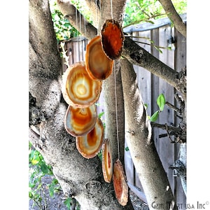 May include: A hanging wind chime made of polished agate slices. The slices are various shades of brown, orange, and cream, with concentric patterns. The chime hangs from thin, clear strings against a backdrop of tree branches and a wooden fence.