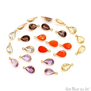 Könnte beinhalten: Eine Sammlung tropfenförmiger Edelsteinanhänger in verschiedenen Farben, darunter transparent, lila, orange und gelb. Jeder Anhänger hat eine goldfarbene Metalleinfassung und eine kleine Schlaufe zum Befestigen an einer Kette. Die Anhänger sind auf weißem Hintergrund angeordnet.