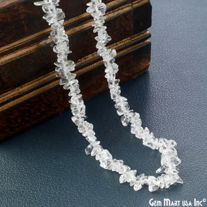 May include: A strand of clear quartz chip beads, strung on a thin cord. The beads are irregular in shape and size, and have a natural, unpolished finish. The beads are displayed against a dark blue background.