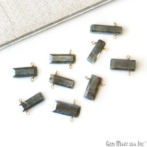 May include: A collection of rectangular, faceted labradorite gemstone beads with copper wire loops for jewelry making. The beads are various shades of gray and blue, scattered on a white surface.