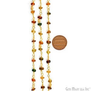 May include: A gold-toned metal chain featuring small, irregularly shaped, multi-coloured gemstone beads in shades of amber, brown, and green, spaced along the links. A copper-coloured one-cent coin is placed next to the chain for scale reference.
