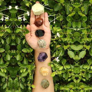 Peut inclure: Une main tenant sept pierres de couleurs différentes, dont du vert, du blanc, du violet, du marron, du noir et du jaune. Les pierres sont disposées en ligne verticale sur la paume de la main.