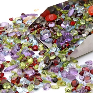 May include: A collection of assorted gemstones in various shapes and sizes, including amethyst, peridot, garnet, and topaz. The gemstones are in shades of purple, green, red, and blue. A metal scoop is partially visible, holding some of the gems.