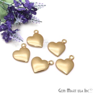Puede incluir: Cinco dijes de corazón de color dorado con un bucle en la parte superior. Los dijes están dispuestos en un semicírculo sobre un fondo blanco.
