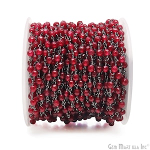 May include: A spool of faceted, deep red gemstone beads strung on a dark metal chain. The beads are a vibrant, saturated color, and the chain is tightly wound on a clear plastic spool. The beads are small and round, and the chain is delicate.