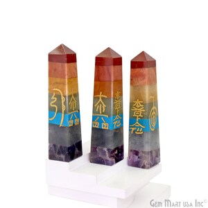 Pode incluir: Três obeliscos de pedras preciosas coloridas com camadas de vermelho, laranja, amarelo, verde, azul e roxo. Cada obelisco apresenta símbolos dourados e repousa sobre uma plataforma branca escalonada. As pedras são polidas.