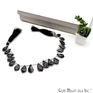 May include: A strand of black and white snowflake obsidian gemstone beads, each bead is faceted and shaped like a teardrop. The beads are strung on silver wire and have black tassels on each end.