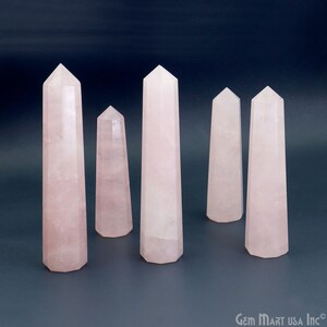 Pode incluir: Cinco pontas de cristal de quartzo rosa, também conhecidas como varinhas, estão dispostas sobre uma superfície cinza. As pontas são de vários tamanhos e formas.