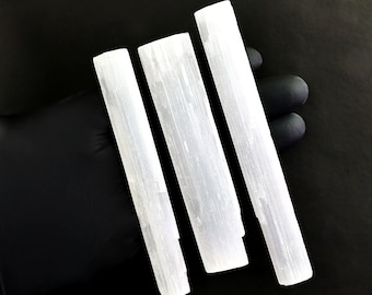 May include: Three white selenite wands, also known as selenite sticks, are arranged vertically on a black surface. The wands are smooth and have a natural, striated texture.
