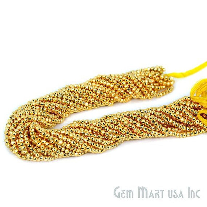May include: Close-up of two strands of gold-colored beads. The beads are small, round, and tightly strung together, creating a textured surface. The strands are arranged in a curved shape against a white background. The ends of the strands have yellow tassels.