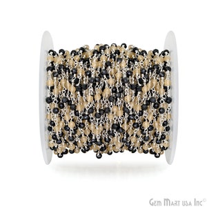 Pode incluir: Um carretel de contas de pedras preciosas pretas e bege enfiadas em uma corrente de prata. As contas são facetadas e têm um acabamento polido.