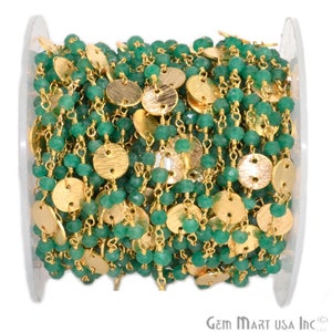 May include: A spool of beaded chain featuring small, round, faceted green beads and gold-colored, flat, circular accents. The chain is wound on a clear plastic spool, ready for use in jewelry making or craft projects.