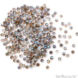 May include: A collection of small, round labradorite gemstones. The stones display a mix of colors, including blues, greens, and golds, with a shimmering, iridescent effect. The gemstones are scattered across a white surface.